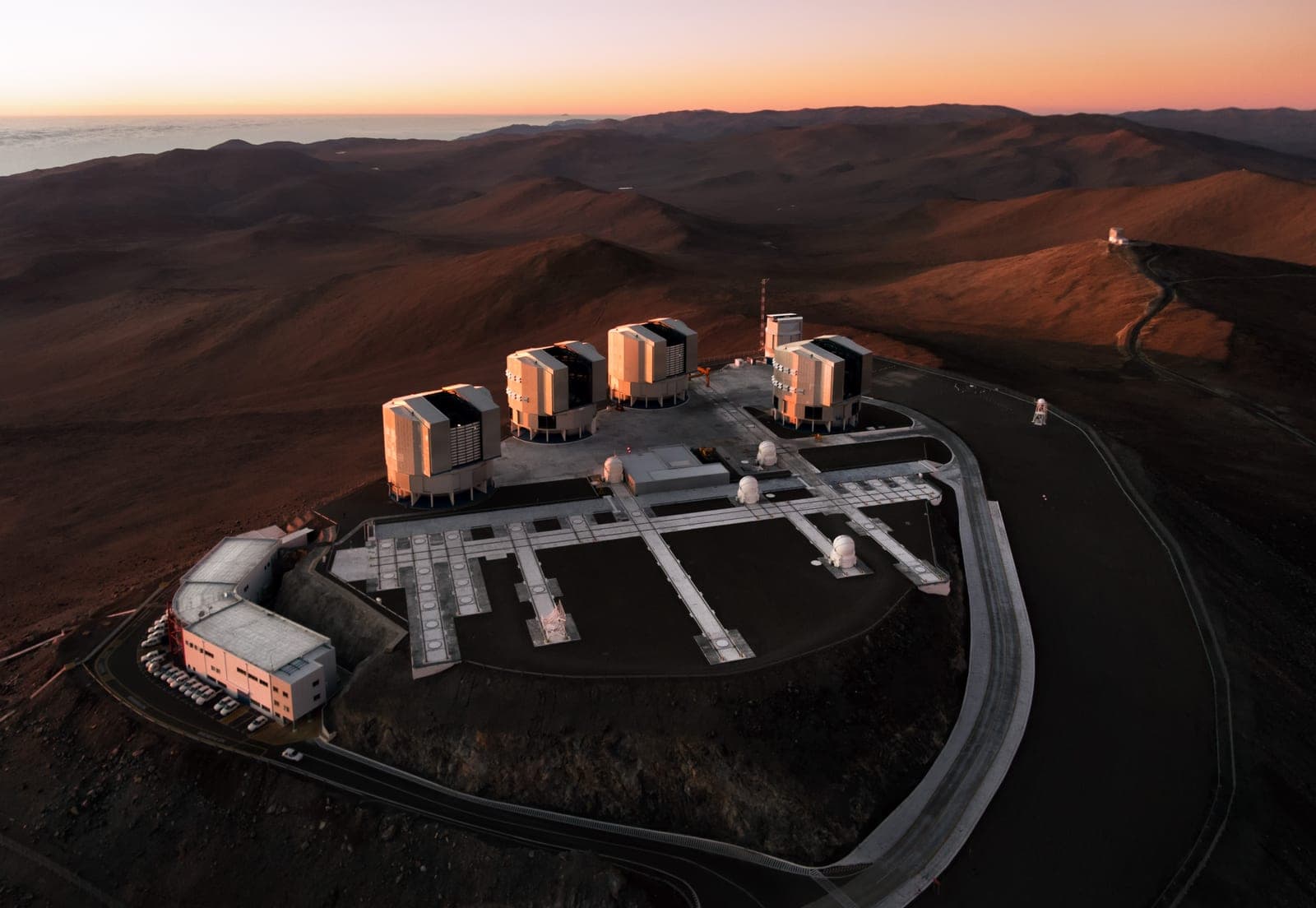 Four large white telescope domes on a flattened mountain top in a red-brown desert at sunset.