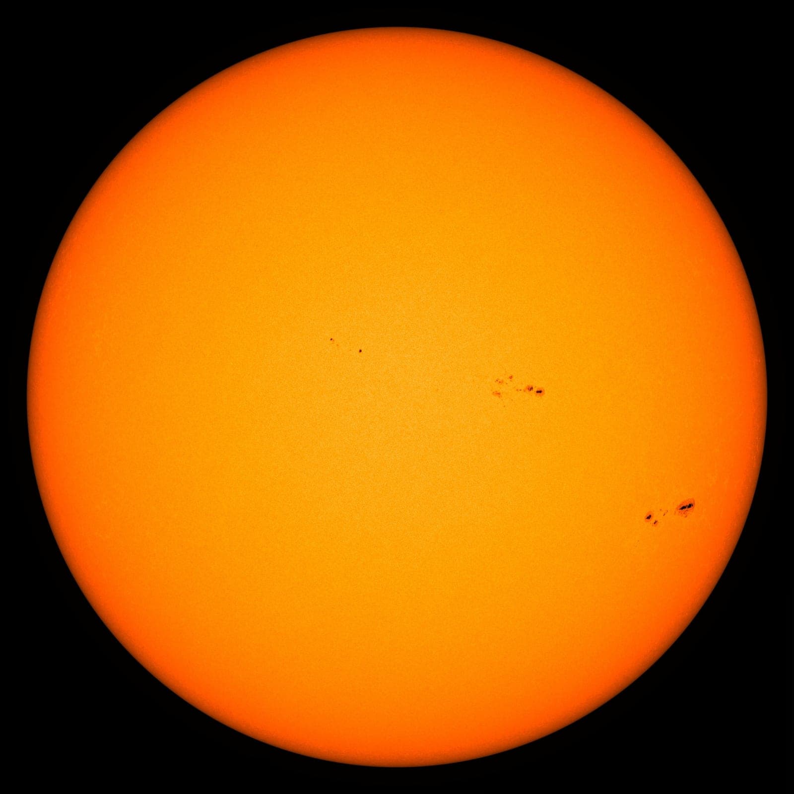A smooth orange disk of the Sun with a few small dark sunspots.